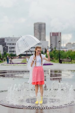 Sarı çizmeli ve fıskiyenin içinde şeffaf şemsiyeli iki örgülü genç ve güzel kız..