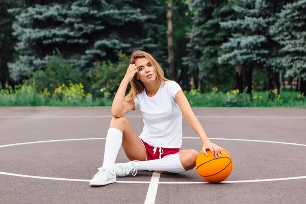 Menina bonita vestida com t-shirt branca, shorts e tênis, senta-se em um campo de basquete com bola . — Fotografia de Stock