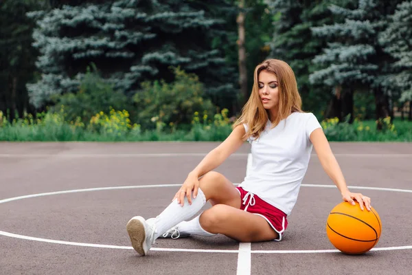 Hermosa joven vestida con camiseta blanca, pantalones cortos y zapatillas de deporte, se sienta en una cancha de baloncesto con pelota . —  Fotos de Stock