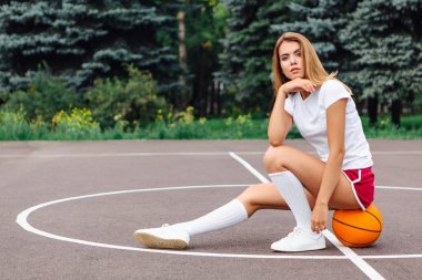 Beyaz tişört, şort ve spor ayakkabıları giymiş güzel genç kız, topa bir basketbol sahası oturur.