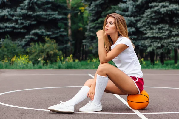Bella ragazza vestita con t-shirt bianca, pantaloncini e scarpe da ginnastica, si siede su un campo da basket sulla palla . — Foto Stock