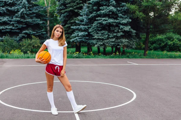 Όμορφη νεαρή κοπέλα ντυμένη με λευκό μπλουζάκι, σορτσάκι και πάνινα παπούτσια, παίζει με μια μπάλα σε ένα γήπεδο μπάσκετ. — Φωτογραφία Αρχείου