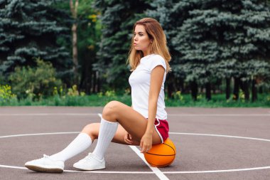 Beyaz tişört, şort ve spor ayakkabıları giymiş güzel genç kız, topa bir basketbol sahası oturur.