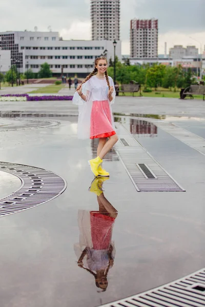 Gadis cantik basah muda dengan dua kepang sepatu bot kuning berdiri di dekat air mancur. Gadis muda mencerminkan di tanah basah — Stok Foto