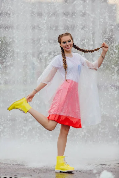 Joven chica bonita mojada con dos trenzas en botas amarillas y con paraguas transparente en el interior de la fuente . — Foto de Stock