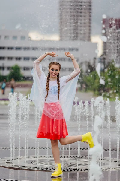 Gadis cantik basah muda dengan dua kepang sepatu bot kuning dan dengan payung transparan berdiri di dalam air mancur . — Stok Foto