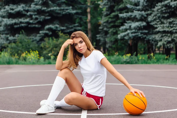 Frumoasă fată tânără îmbrăcată în tricou alb, pantaloni scurți și adidași, stă pe un teren de baschet cu minge . — Fotografie, imagine de stoc