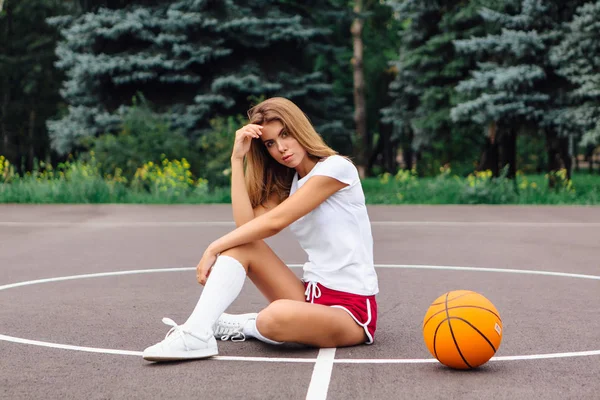 Mooi jong meisje gekleed in wit t-shirt, shorts en sneakers, zit op een basketbalveld met bal. — Stockfoto