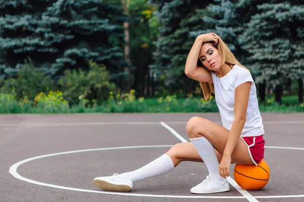 Bella ragazza vestita con t-shirt bianca, pantaloncini e scarpe da ginnastica, si siede su un campo da basket sulla palla . — Foto Stock