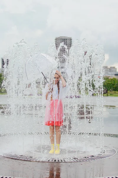 Giovane ragazza bella bagnata con due trecce in stivali gialli e con portaombrelli trasparente all'interno della fontana . — Foto Stock