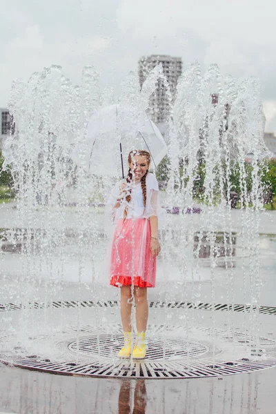 Sarı çizmeli ve fıskiyenin içinde şeffaf şemsiyeli iki örgülü genç ve güzel kız..
