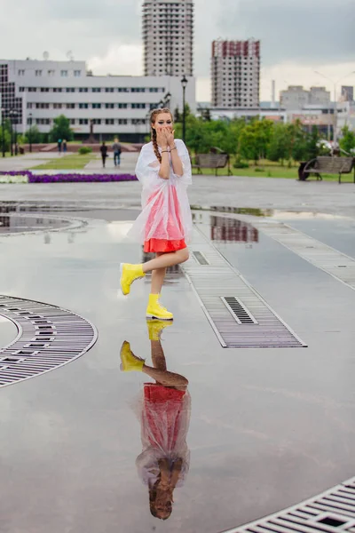 Menina bonita molhada nova com duas tranças em botas amarelas fica perto da fonte. Menina refletindo no chão molhado — Fotografia de Stock