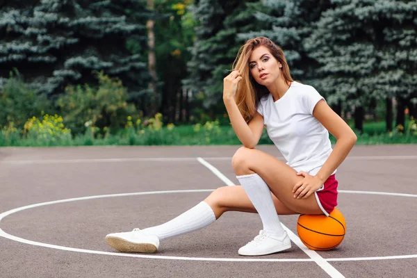 Menina bonita vestida com t-shirt branca, shorts e tênis, senta-se em um campo de basquete na bola . — Fotografia de Stock