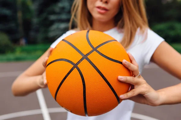 Beautiful young girl dressed in white t-shirt, shorts and sneakers, plays with a ball on a basketball court.