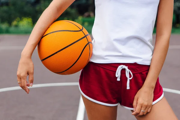 Wanita muda mengenakan kaos putih, celana pendek memegang bola di lapangan basket . — Stok Foto