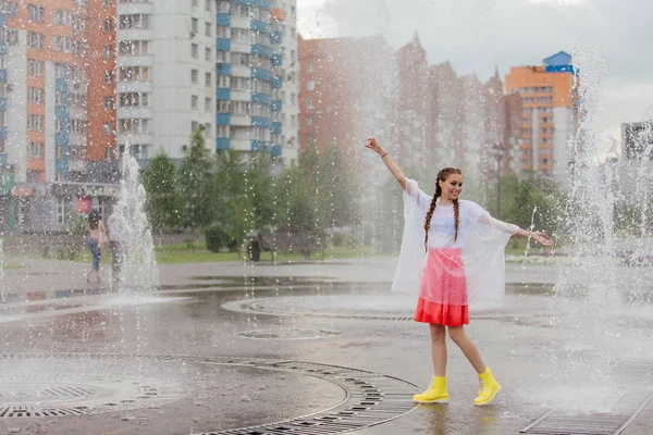 Молодая мокрая красивая девушка с двумя косичками в жёлтых сапогах стоит у фонтана . — стоковое фото