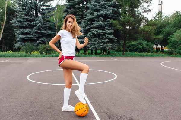 Vacker ung flicka klädd i vit t-shirt, shorts och sneakers, spelar med en boll på en basketplan. — Stockfoto
