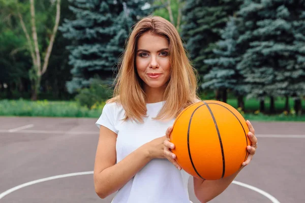 Hermosa joven vestida con camiseta blanca, pantalones cortos y zapatillas de deporte, juega con una pelota en una cancha de baloncesto . — Foto de Stock
