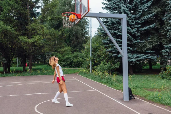 Gadis muda yang cantik mengenakan kaos putih, celana pendek dan sepatu olahraga, bermain dengan bola di lapangan basket . — Stok Foto