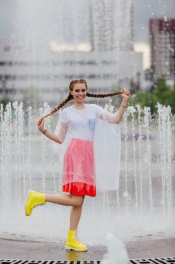 Sarı çizmeli ve fıskiyenin içinde şeffaf şemsiyeli iki örgülü genç ve güzel kız..