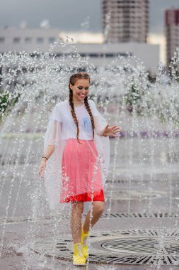 Sarı çizmeli ve fıskiyenin içinde şeffaf şemsiyeli iki örgülü genç ve güzel kız..