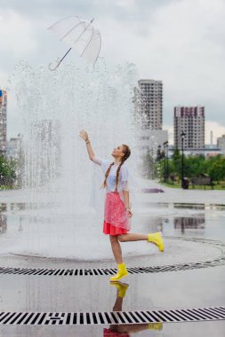 Genç güzel kız atma ve şeffaf şemsiye çeşme yakalamak sarı Çizmeli iki örgüler ile.