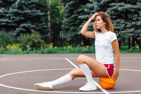 Красивая девушка, одетая в белую футболку, шорты и кроссовки, сидит на баскетбольной площадке на мяче . — стоковое фото