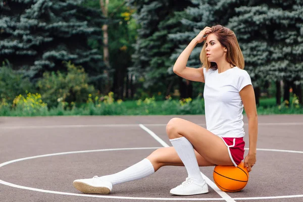 Bella ragazza vestita con t-shirt bianca, pantaloncini e scarpe da ginnastica, si siede su un campo da basket sulla palla . — Foto Stock