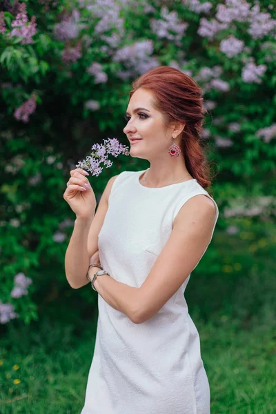 Charming woman wearing beautiful white dress standing next to lilac bush — Stock Photo, Image
