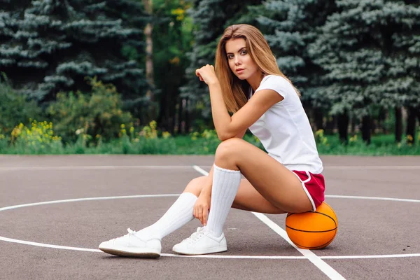 Mooi jong meisje gekleed in wit t-shirt, shorts en sneakers, zit op een basketbalveld op de bal. — Stockfoto