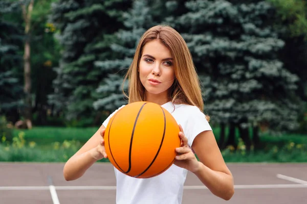 Menina bonita vestida com t-shirt branca, shorts e tênis, joga com uma bola em um campo de basquete . — Fotografia de Stock