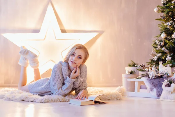 Schöne junge Frau legt und liest Buch unter dem Weihnachtsbaum in einem Urlaub Interieur — Stockfoto