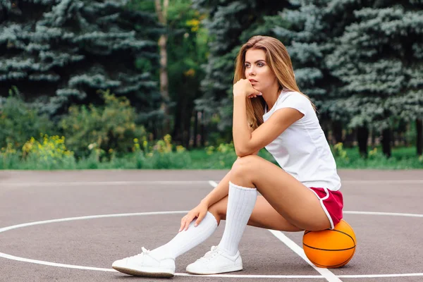 Bella ragazza vestita con t-shirt bianca, pantaloncini e scarpe da ginnastica, si siede su un campo da basket sulla palla . — Foto Stock