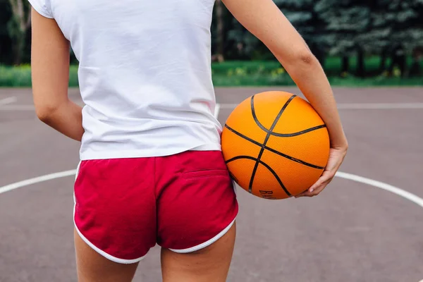 Young woman dressed in white t-shirt, shorts holding a ball on a basketball court.