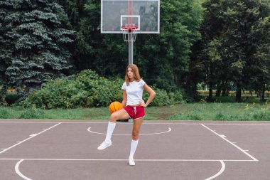 Güzel genç kız beyaz tişört, şort ve spor ayakkabısı, giyinmiş bir basketbol sahası üzerinde bir top ile çalış.