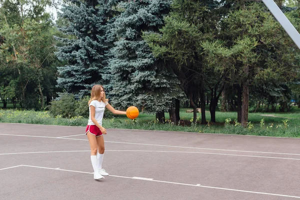 Menina bonita vestida com t-shirt branca, shorts e tênis, joga com uma bola em um campo de basquete . — Fotografia de Stock