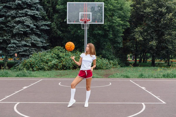 सुंदर युवा लड़की सफेद टी-शर्ट, शॉर्ट्स और स्नीकर्स में पोशाक, बास्केटबॉल कोर्ट पर एक गेंद के साथ खेलती है . — स्टॉक फ़ोटो, इमेज