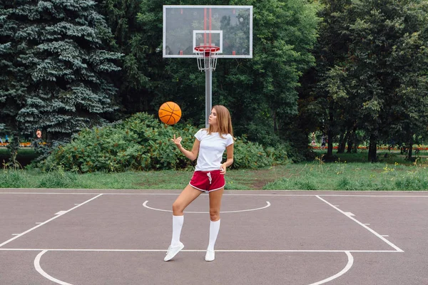 Gadis muda yang cantik mengenakan kaos putih, celana pendek dan sepatu olahraga, bermain dengan bola di lapangan basket . — Stok Foto