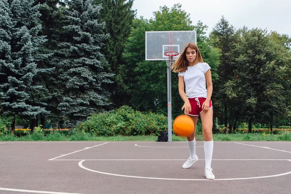 Frumoasă fată tânără îmbrăcată în tricou alb, pantaloni scurți și adidași, se joacă cu o minge pe un teren de baschet . — Fotografie, imagine de stoc