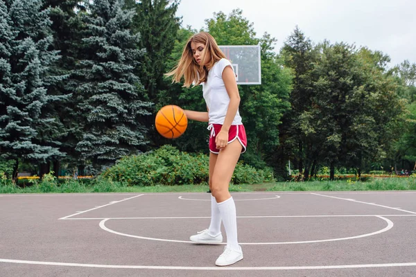 Gadis muda yang cantik mengenakan kaos putih, celana pendek dan sepatu olahraga, bermain dengan bola di lapangan basket . — Stok Foto