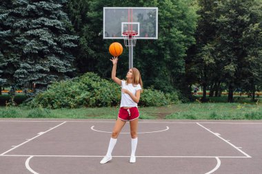 Güzel genç kız beyaz tişört, şort ve spor ayakkabısı, giyinmiş bir basketbol sahası üzerinde bir top ile çalış.