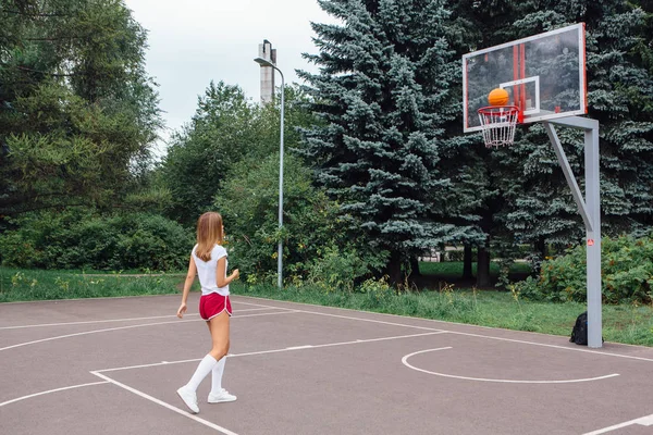 Gadis muda yang cantik mengenakan kaos putih, celana pendek dan sepatu olahraga, bermain dengan bola di lapangan basket . — Stok Foto