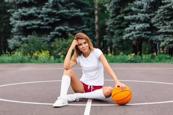 Hermosa joven vestida con camiseta blanca, pantalones cortos y zapatillas de deporte, se sienta en una cancha de baloncesto con pelota . — Foto de Stock