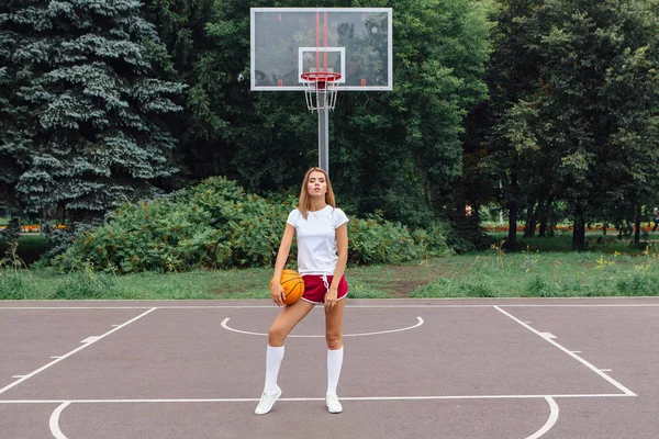 Beautiful young girl dressed in white t-shirt, shorts and sneakers, plays with a ball on a basketball court.