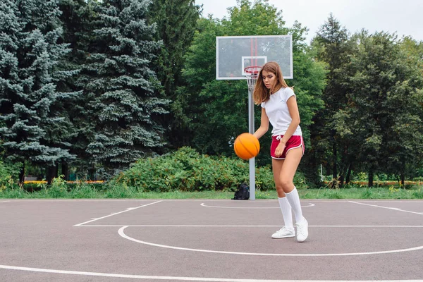 Hermosa joven vestida con camiseta blanca, pantalones cortos y zapatillas de deporte, juega con una pelota en una cancha de baloncesto . — Foto de Stock