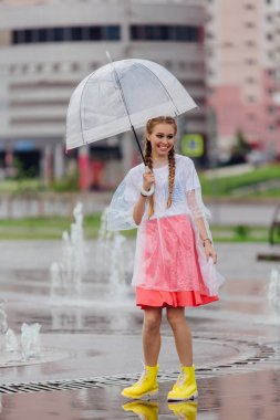 Genç güzel kız sarı çizmeli iki örgüler ve şeffaf şemsiye standları Çeşme yakınındaki.
