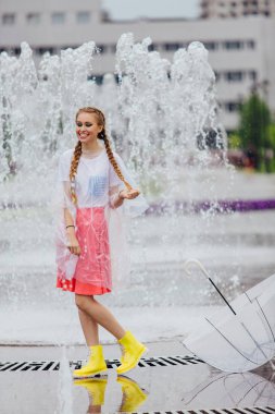 Genç güzel kız sarı çizmeli iki örgüler ve şeffaf şemsiye standları Çeşme yakınındaki.