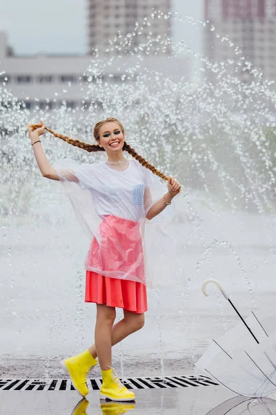 Joven chica bonita con dos trenzas en botas amarillas y con paraguas transparente se levanta cerca de la fuente . — Foto de Stock