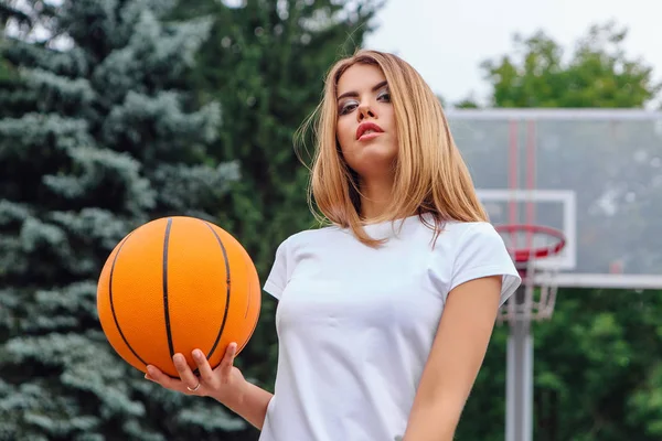 Schönes junges Mädchen in weißem T-Shirt, kurzer Hose und Turnschuhen, spielt mit einem Ball auf einem Basketballfeld. — Stockfoto