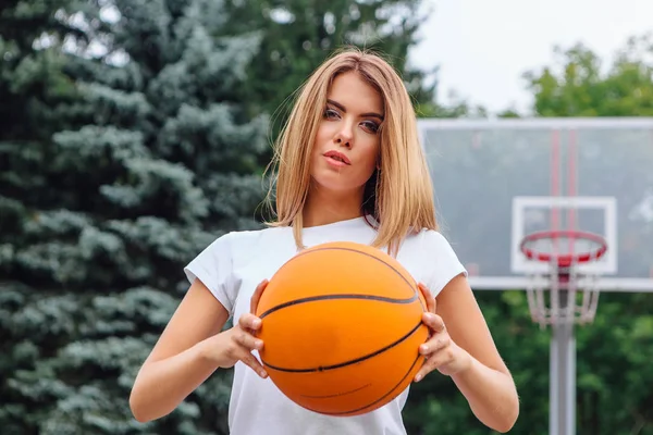 Gadis muda yang cantik mengenakan kaos putih, celana pendek dan sepatu olahraga, bermain dengan bola di lapangan basket . — Stok Foto